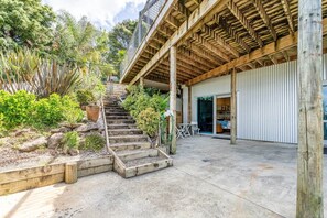 A wooden staircase leading to a covered outdoor area with a metal railing, surrounded by greenery and a building with a corrugated metal exterior. - Sandy Soul Beach Studio (Whitianga)