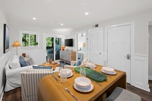 A modern dining area with a wooden table set for four, a sofa, and a television in the background. - The Tidal Cottage - Beach Access + Pools (Hilton Head Island)