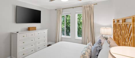 Un dormitorio con cama, cómoda, televisor, ventilador de techo y ventana con cortinas.
