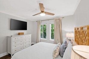 Quarto com cama, cômoda, televisão, ventilador de teto e janela com cortinas.