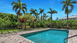 A swimming pool with a clear blue water surface, surrounded by a paved deck and lush greenery including palm trees and flowering plants. - Luxury Downtown Retreat | Steps to 5th Ave & Beach (Naples)