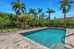 A swimming pool with a clear blue water surface, surrounded by a paved deck and lush greenery including palm trees and flowering plants. - Luxury Downtown Retreat | Steps to 5th Ave & Beach (Naples)