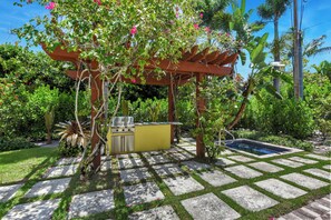 Un espace cuisine extérieur doté d’un barbecue et d’un évier, entouré d’une végétation luxuriante et agrémenté d’une pergola.