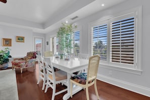A dining area with a white table and chairs, a potted plant, and a view through large windows with blinds. - Luxury Downtown Retreat | Steps to 5th Ave & Beach (Naples)