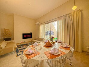 A modern living room with a dining area, a flat-screen TV, and a large window with curtains. - The Blue Room at Riad Timija: En-suite, Breakfast & Rooftop in the Medina (Marrakesh)