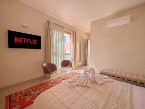 A hotel room with a bed, a TV mounted on the wall, a window with curtains, a small table, two chairs, and a decorative swan on the bed. - The Blue Room at Riad Timija: En-suite, Breakfast & Rooftop in the Medina (Marrakesh)