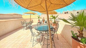 An outdoor patio with a round table and chairs, a large woven umbrella, and potted plants. - The Blue Room at Riad Timija: En-suite, Breakfast & Rooftop in the Medina (Marrakesh)