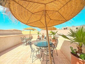 An outdoor patio with a round table and chairs, a large woven umbrella, and potted plants. - The Blue Room at Riad Timija: En-suite, Breakfast & Rooftop in the Medina (Marrakesh)
