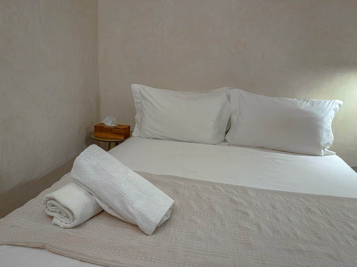 Un lit bien fait, avec du linge de lit blanc et des oreillers, une serviette blanche roulée posée sur le lit, et une petite table de chevet en bois sur laquelle se trouve une boîte de mouchoirs.