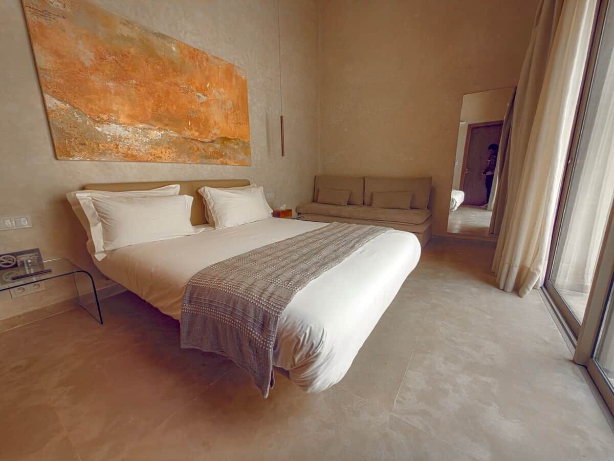 A bedroom with a large bed, a bedside table, a nightstand, a window with curtains, and a painting on the wall.