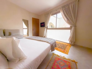 A hotel room with a bed, a TV, a window with curtains, and a patterned rug. - The White Room at Riad Timija: En-suite, Breakfast & Rooftop in the Medina (Marrakesh)