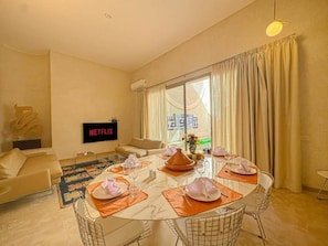 A modern living room with a dining area, a flat-screen TV, and a large window with curtains. - The Red Room at Riad Timija: En-suite, Breakfast & Rooftop in the Medina (Marrakesh)