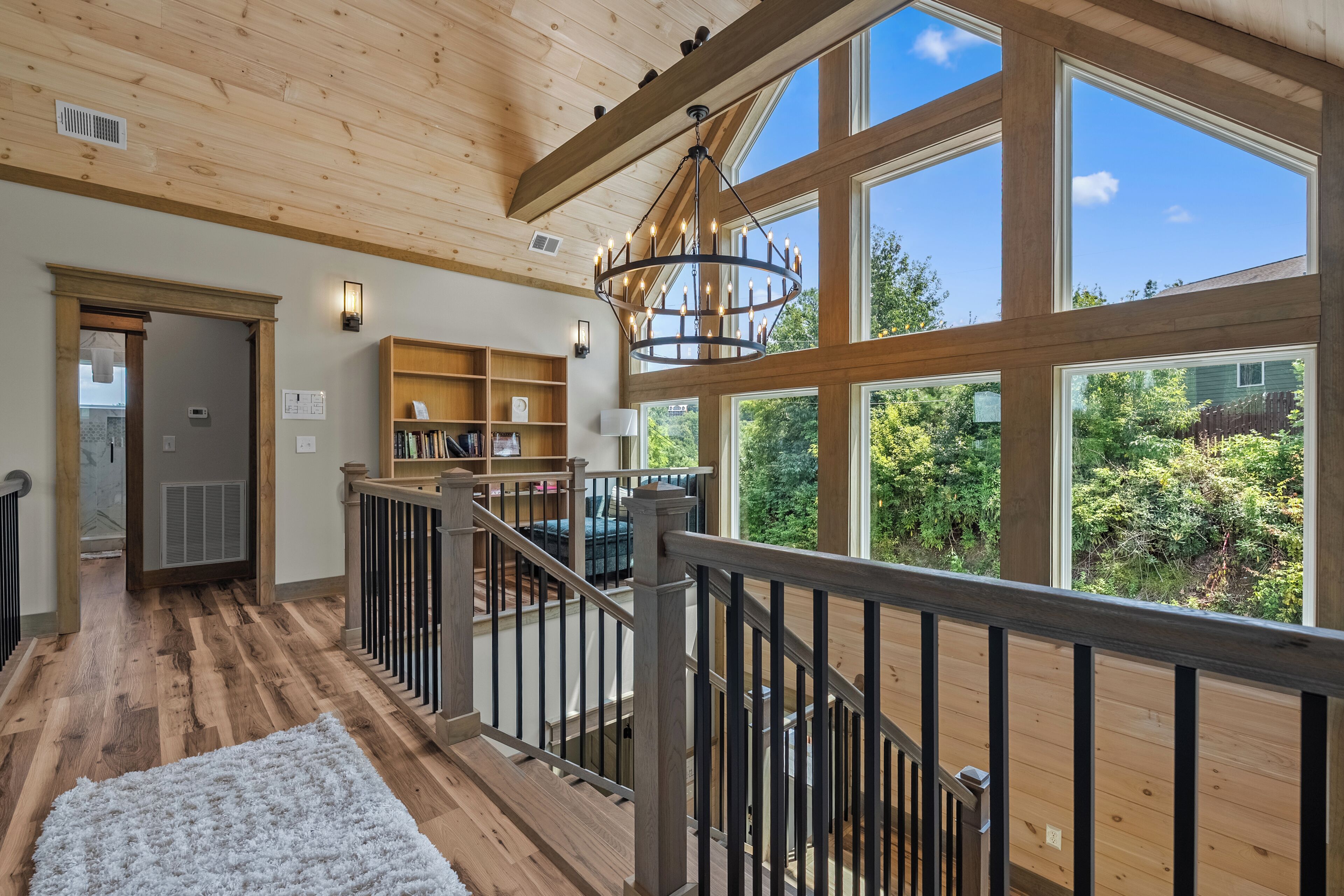 A spacious room with a wooden floor, a staircase with a railing, and large windows offering a view of greenery outside.