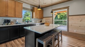 A modern kitchen with wooden cabinets, a large island with stools, pendant lighting, and a view of greenery outside. - Gatlinburg Getaway with Hot Tub, Mountain Views, Theater, Game Room & Smoky Mountains (Gatlinburg)