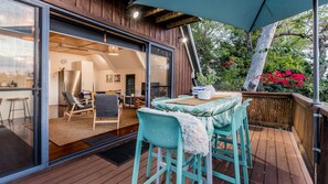 A wooden deck with outdoor seating, a glass door leading to an interior space, and a view of a garden with red flowers. - Riverbank 1770  waterfront (Seventeen Seventy)