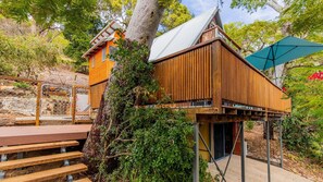 Une cabane en bois perchée dans un arbre, accessible par un escalier en métal, et entourée de verdure.