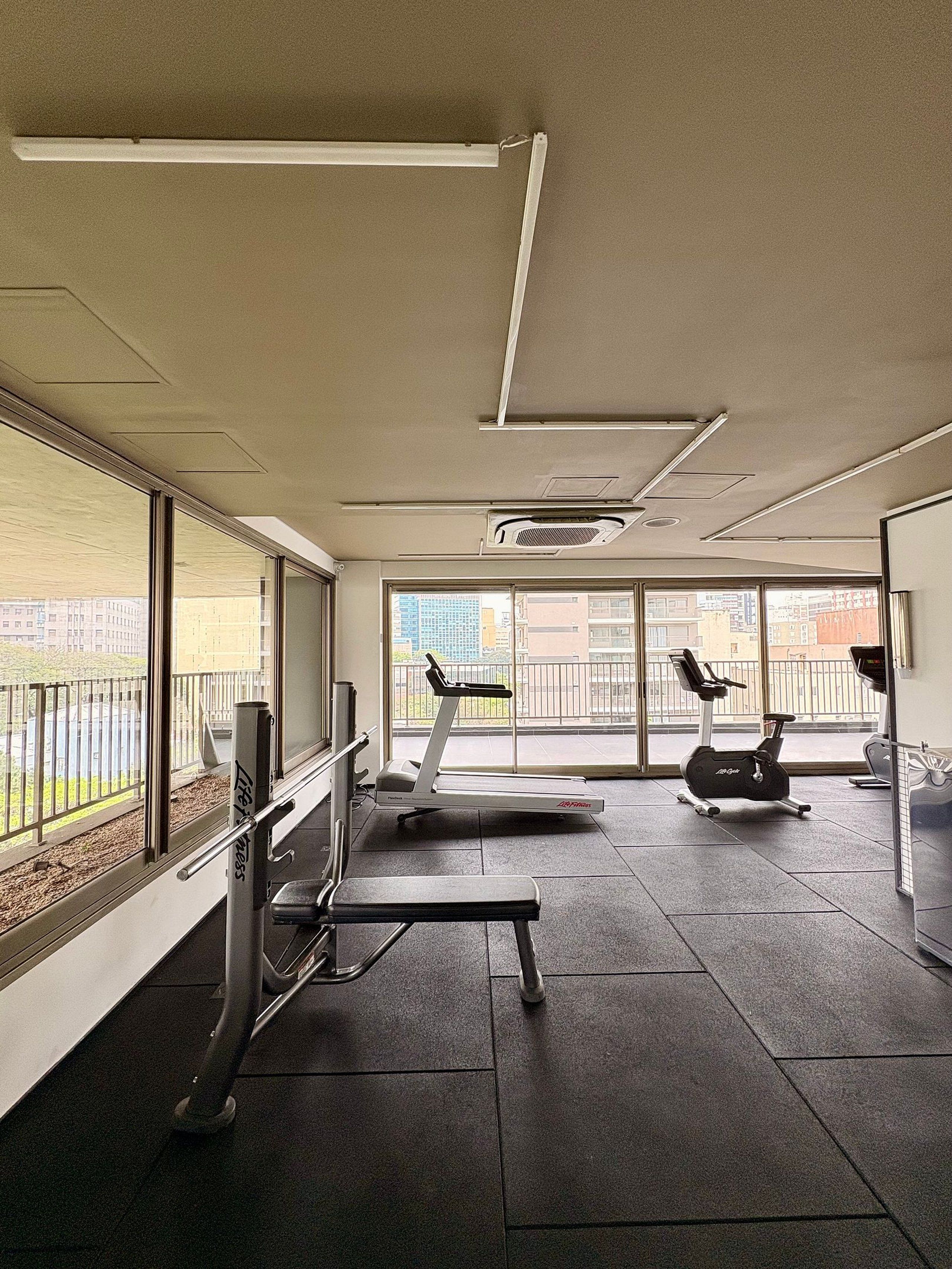 Une salle de sport équipée de matériel d’exercice, dotée de grandes fenêtres et offrant une vue sur la ville.