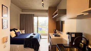 Habitación de hotel compacta con cama, kitchenette, baño pequeño y vistas a la ciudad desde la ventana.