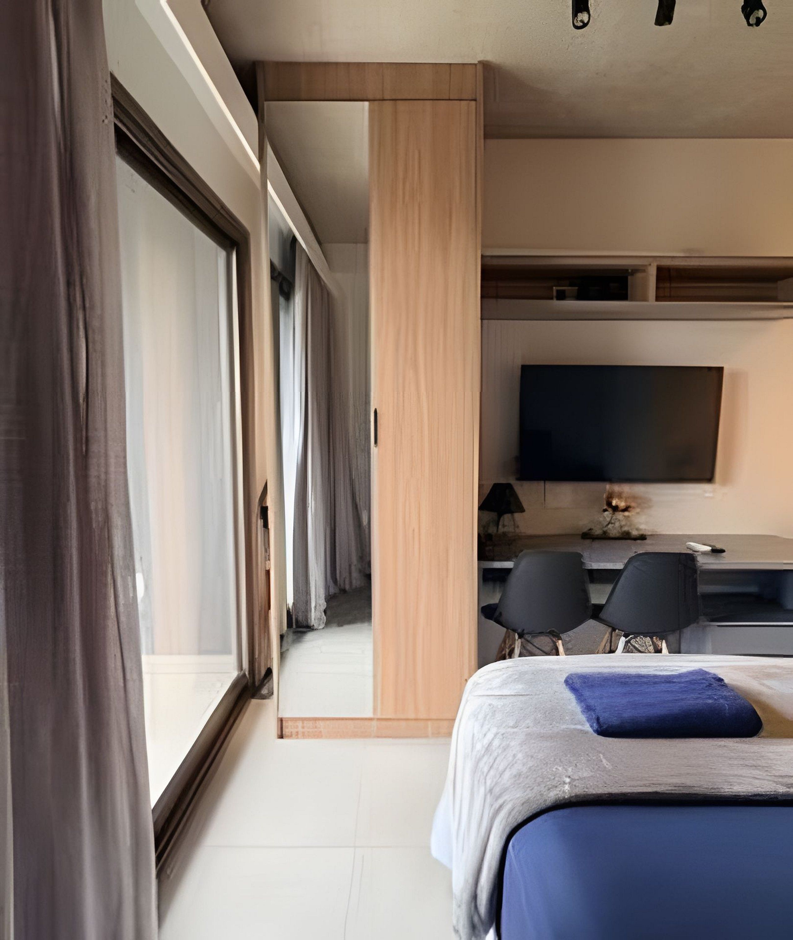 A modern hotel room with a large bed, a desk with two chairs, a television, and a sliding glass door leading to a balcony.