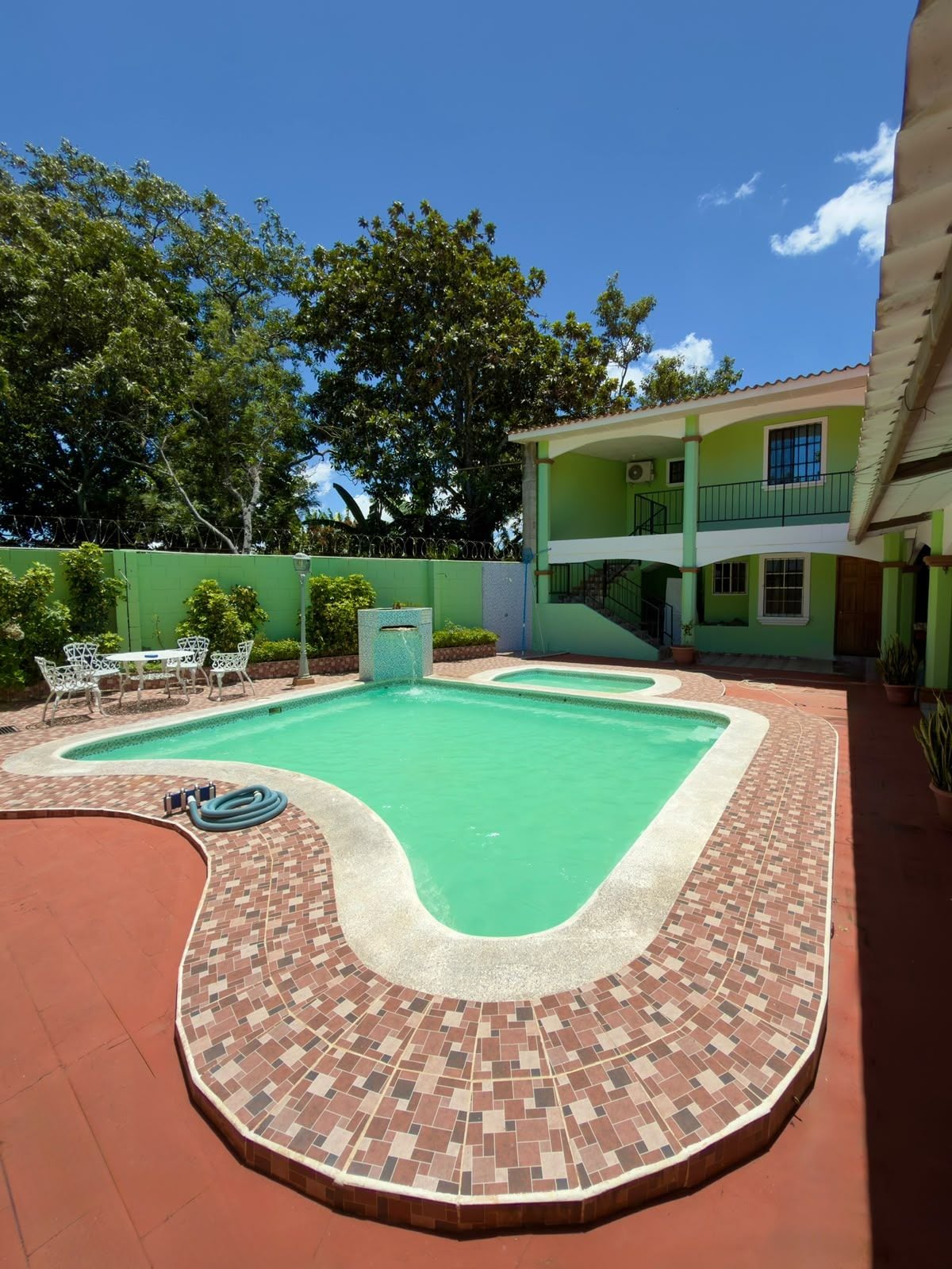 A swimming pool with a tiled deck, a covered seating area, and a two-story building in the background.