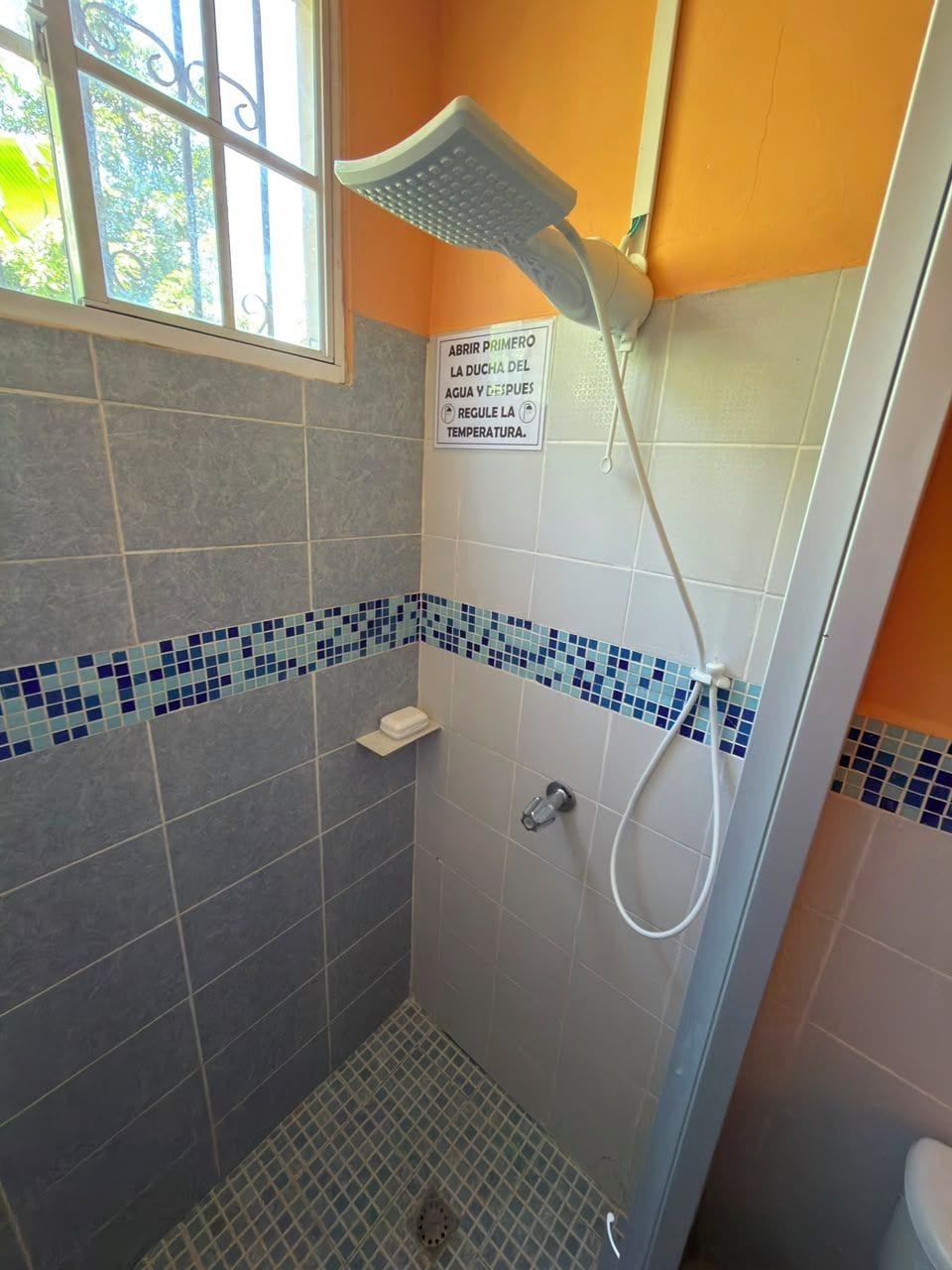 A shower with a handheld showerhead, a tiled floor, and a sign instructing to open the shower door first.