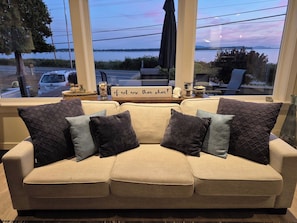 En beige sofa med flere puder, et træsidebord med dekorative genstande og udsigt over et vandareal gennem store vinduer.