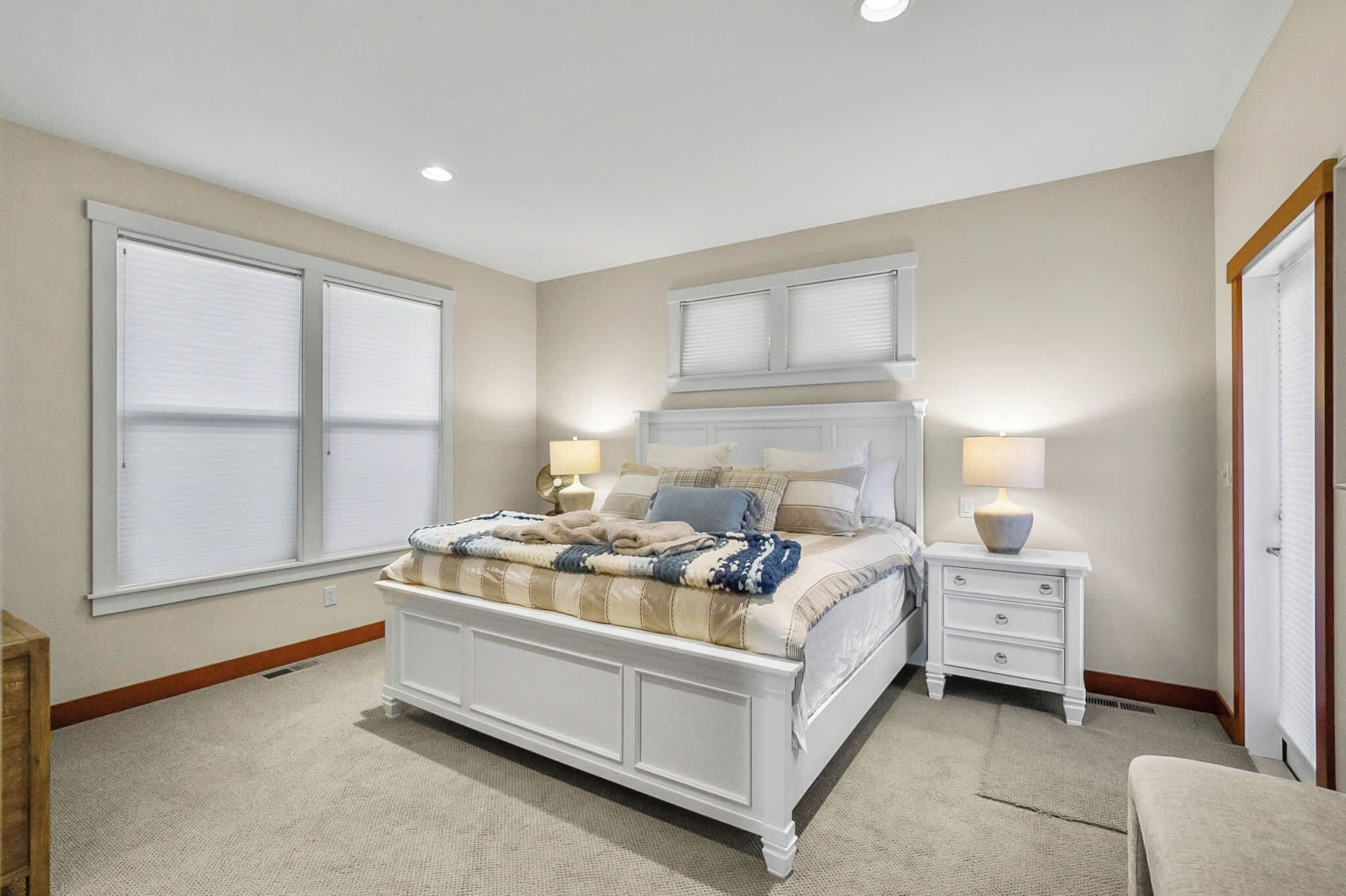 A bedroom with a large bed, two bedside tables, a window with blinds, and a door.