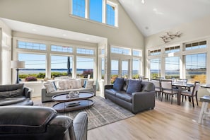 A spacious living room with a large window overlooking a body of water, a dining area with a view, and a high ceiling with skylights.
