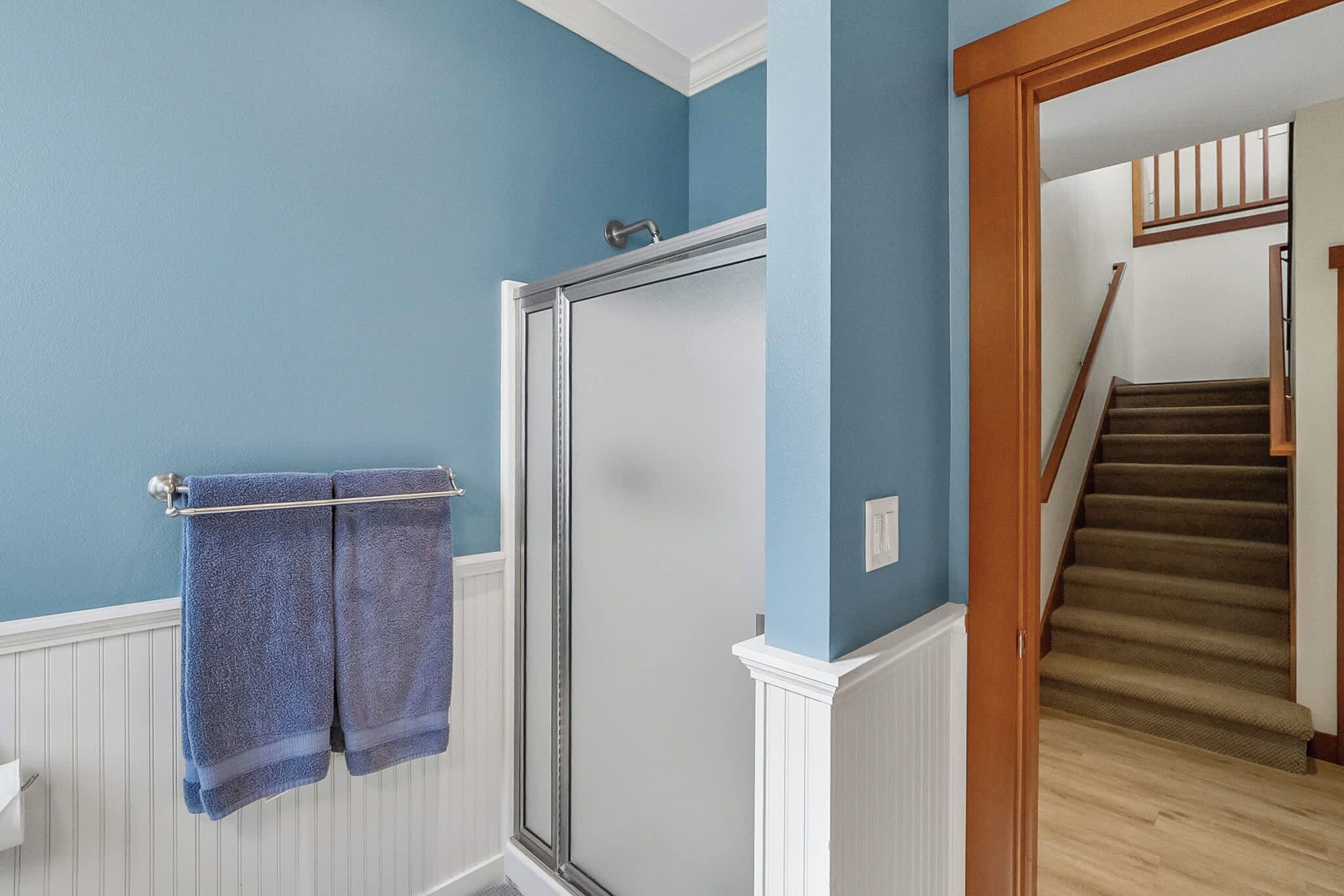 Une salle de bain dotée d’une porte de douche en verre, d’un porte-serviettes avec deux serviettes et d’un escalier en bois menant à l’étage supérieur.