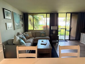 A living room with a sofa, a dining table, chairs, a lamp, and a view of the outdoors through large windows. - Upgraded 1BD/1.5BA ocean view condo steps away from fantastic Kam 1 beach (kihei)