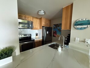 A modern kitchen with wooden cabinets, stainless steel appliances, a marble countertop, and a wall clock. - Upgraded 1BD/1.5BA ocean view condo steps away from fantastic Kam 1 beach (kihei)