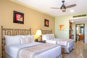 Una habitació d'hotel amb dos llits, ventilador de sostre, obres d'art a les parets i zona de cuina compacta.