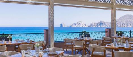 An outdoor dining area with wicker furniture and tables set for guests, overlooking a clear blue sea and rocky coastline.