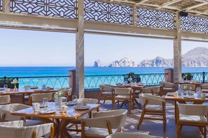An outdoor dining area with wicker furniture and tables set for guests, overlooking a clear blue sea and rocky coastline. - Junior Suite in Cabo San Lucas on Medano Beach with Amazing Views of the Arch! (Cabo San Lucas)