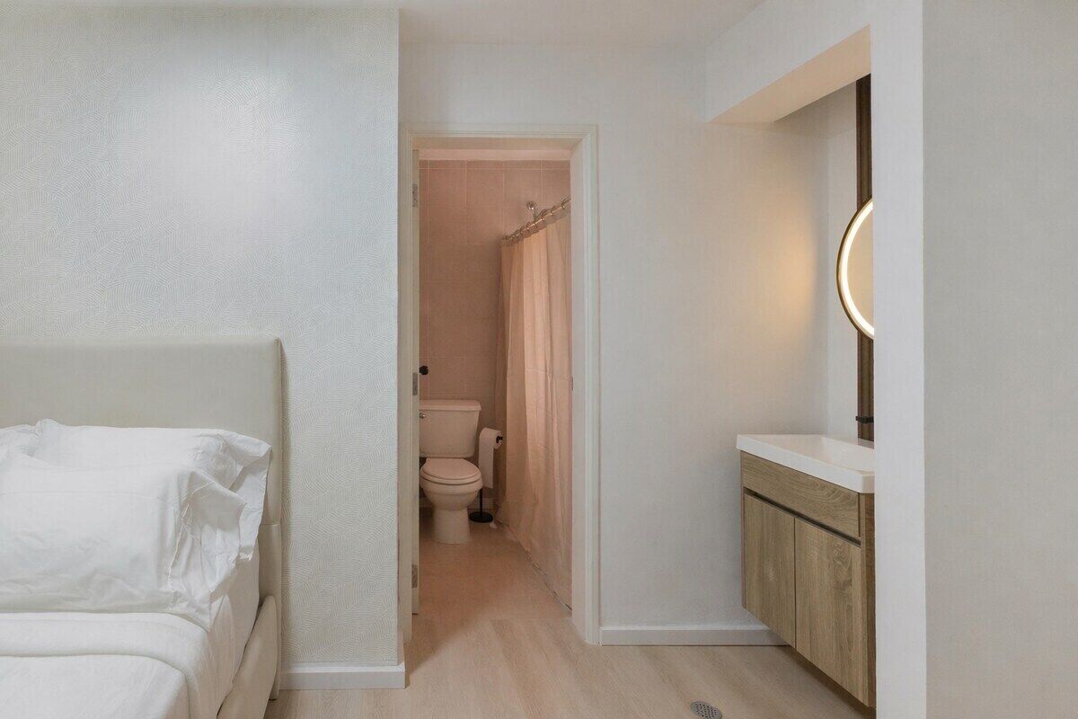 A bedroom with a bed, a bathroom with a toilet and shower curtain, and a vanity with a mirror.