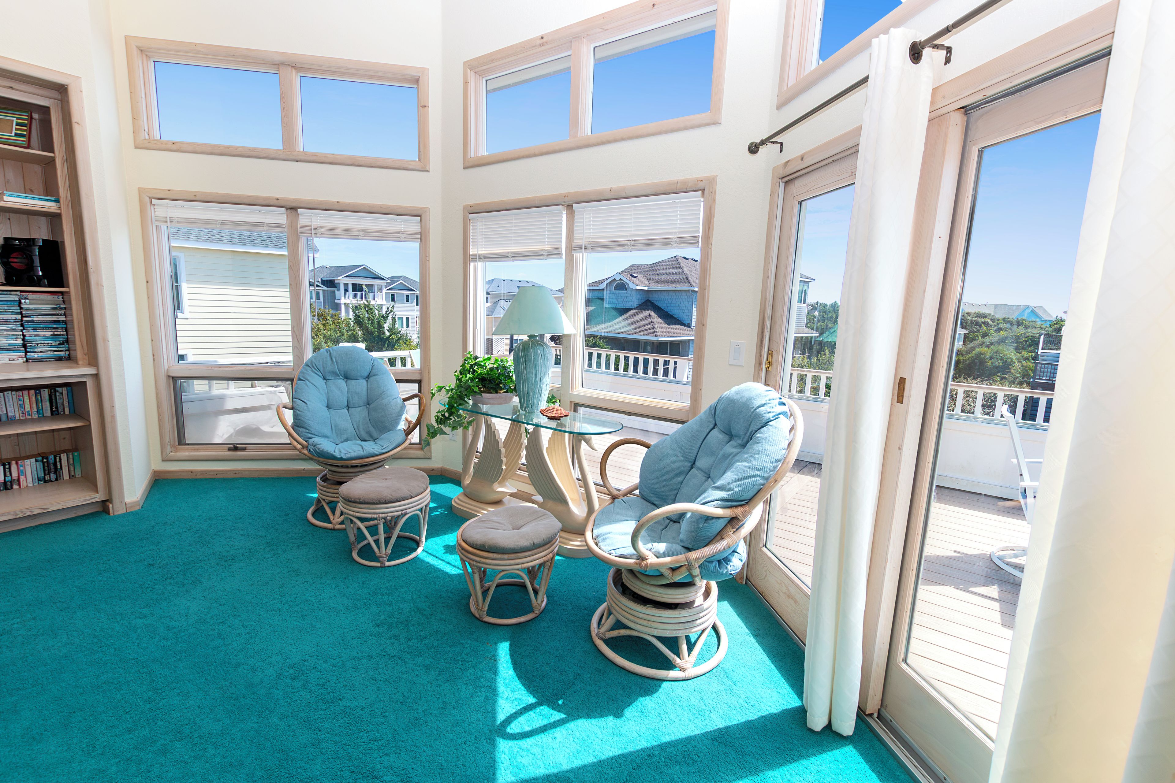 A sunlit room with a glass table, two wicker chairs, a bookshelf, and a view of neighboring houses.