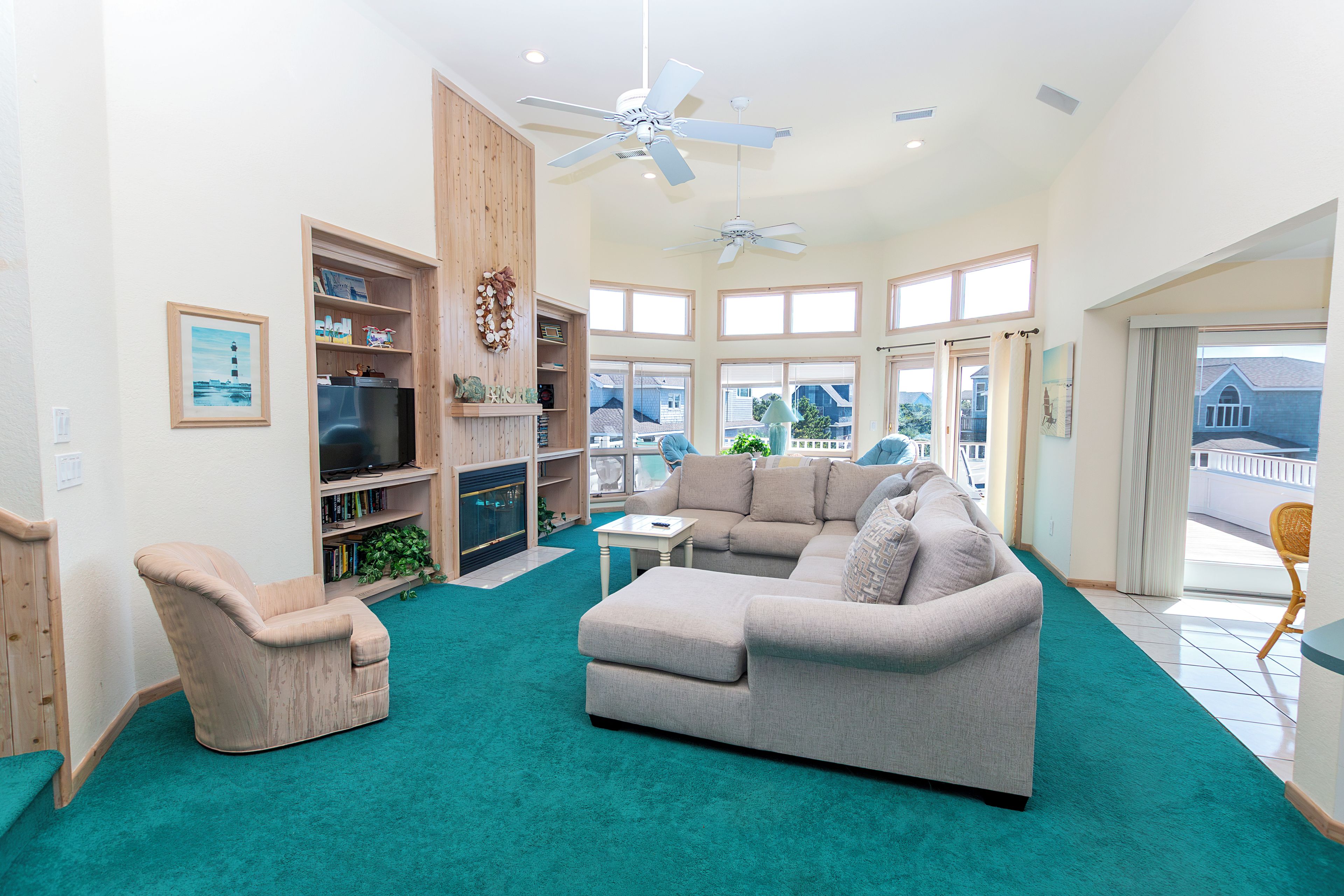A spacious living room with a grey sofa, a beige armchair, and a teal carpet. There are wooden shelves with various items and a fireplace.