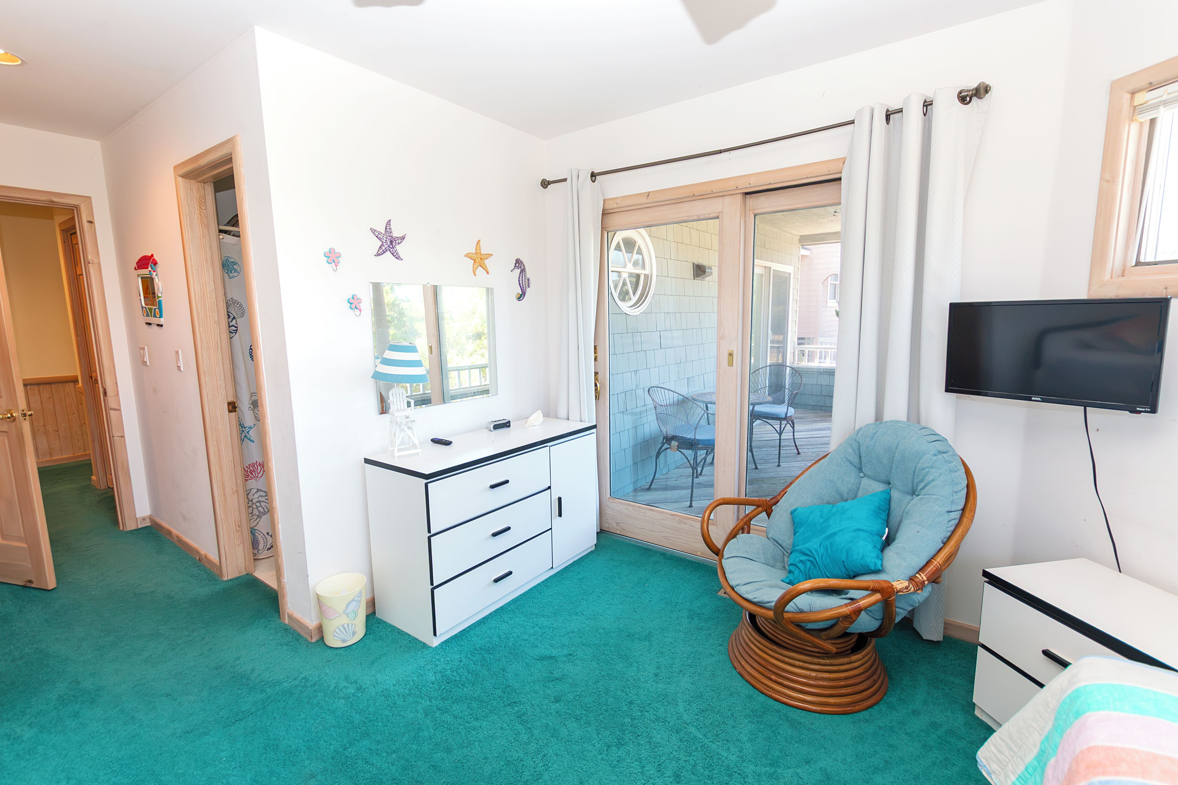 A room with a white dresser, a wicker chair, a television, and a sliding glass door leading to an outdoor area.