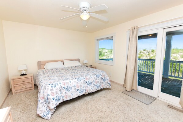 Une chambre à coucher comprenant un lit, une table de chevet, une lampe, un ventilateur de plafond, une fenêtre avec vue et une porte-fenêtre donnant sur un balcon.