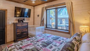 Ein Zimmer mit Holzwandverkleidung, einem Bett, einem Babybett, einer Kommode, einem an der Wand befestigten Fernseher und einem Fenster mit Vorhängen.