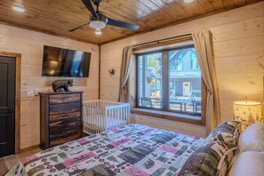 A wooden-paneled room with a bed, a crib, a dresser, a TV mounted on the wall, and a window with curtains. - 2 Cabins - Indoor Pool & 2 Hot Tubs - Sleeps 13 (Pigeon Forge)