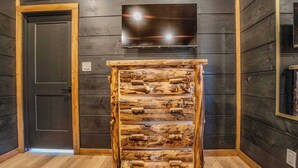 Ein rustikales Holzschränkchen mit mehreren Schubladen vor einer dunkelholzigen Wand, darüber ein Flachbildfernseher montiert und eine Tür links.