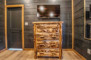 A rustic wooden cabinet with multiple drawers against a dark wooden wall, a flat-screen TV mounted above it, and a door to the left. - 2 Cabins - Indoor Pool & 2 Hot Tubs - Sleeps 13 (Pigeon Forge)
