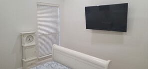 A bedroom with a bed, a white wall-mounted TV, a white clock, and a window with blinds. - Serene 3-bedroom house plus office with AC in Fort Worth (Fort Worth)