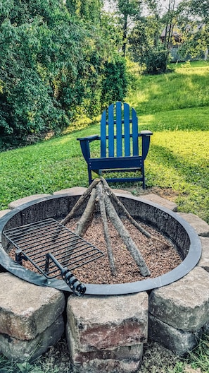 A fire pit with logs and a metal grill, surrounded by stone blocks, with a blue Adirondack chair in the background.