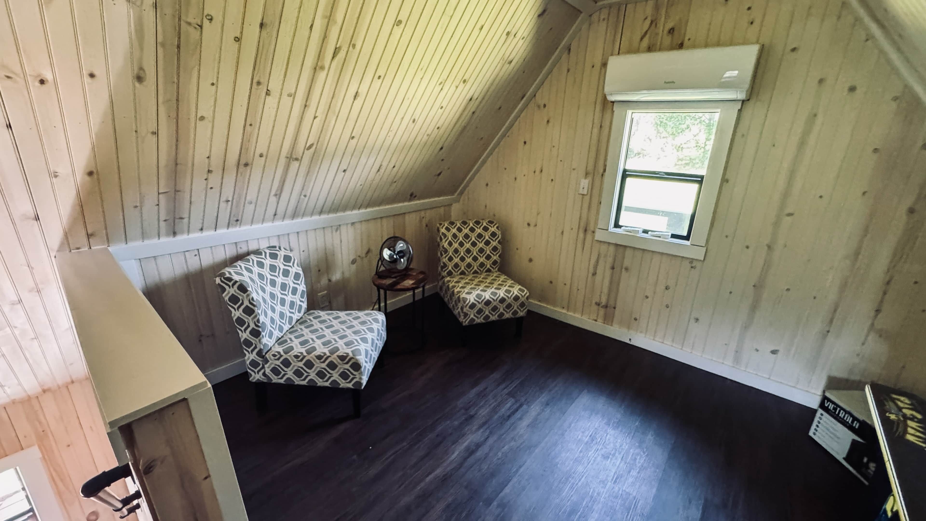 A room with a sloped ceiling, two patterned chairs, a small table, and a window.