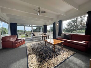 A spacious living room with a large window, a ceiling fan, and a view of the outdoors. - OPPOSITE THE BEACH (Piha)