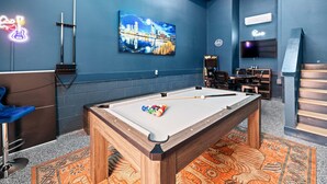 Una mesa de billar con bolas, un letrero de guitarra neón, una pintura de paisaje urbano, un televisor y una zona de bar.