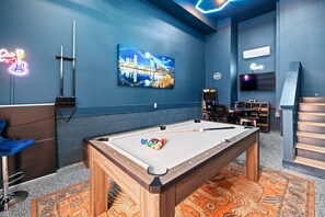 Ein Billardtisch mit Kugeln, ein neonbeleuchtetes Gitarrenschild, ein Stadtbildgemälde, ein Fernseher und ein Barbereich.