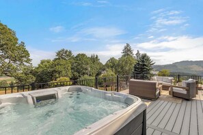 Ein Whirlpool auf einer Holzdielen-Terrasse, mit Gartenmöbeln, umgeben von Bäumen und Blick auf Hügel.